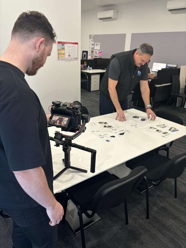 A team member from a social media marketing agency on the Gold Coast and Brisbane filming a professional video shoot indoors, with a man reviewing marketing materials on a table, showcasing behind-the-scenes content creation