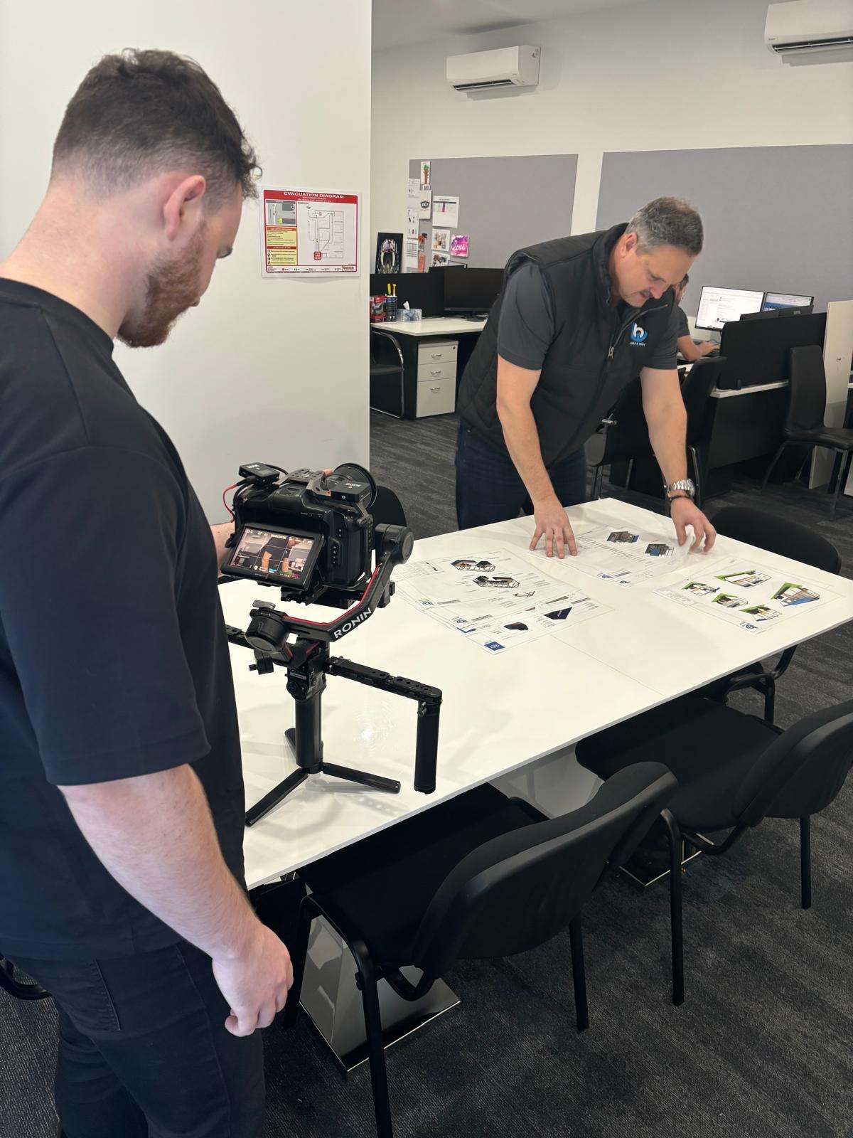 A team member from a social media marketing agency on the Gold Coast and Brisbane filming a professional video shoot indoors, with a man reviewing marketing materials on a table, showcasing behind-the-scenes content creation